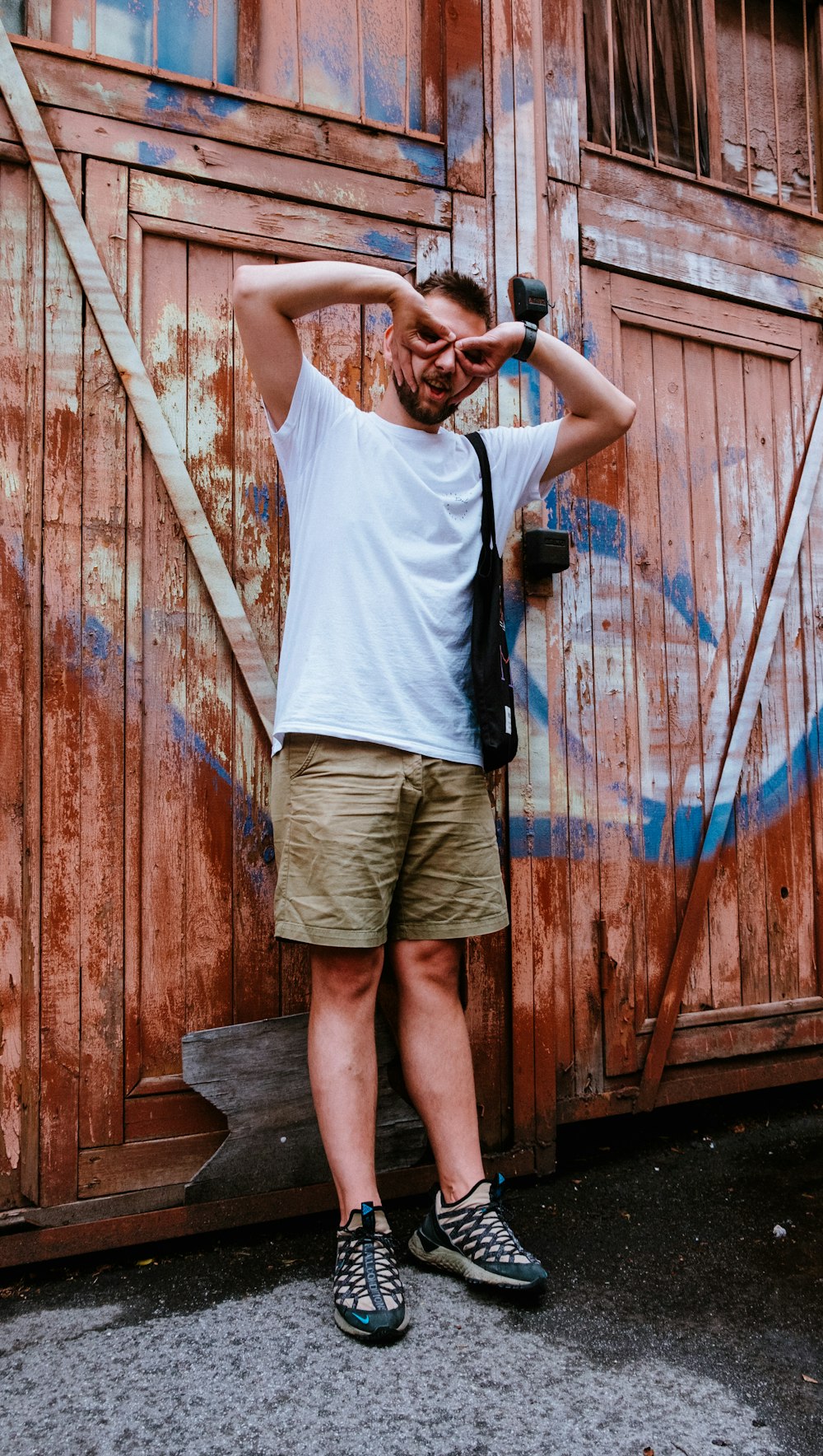 Hombre con camiseta blanca sin mangas y pantalones cortos marrones de pie junto a la pared de madera marrón