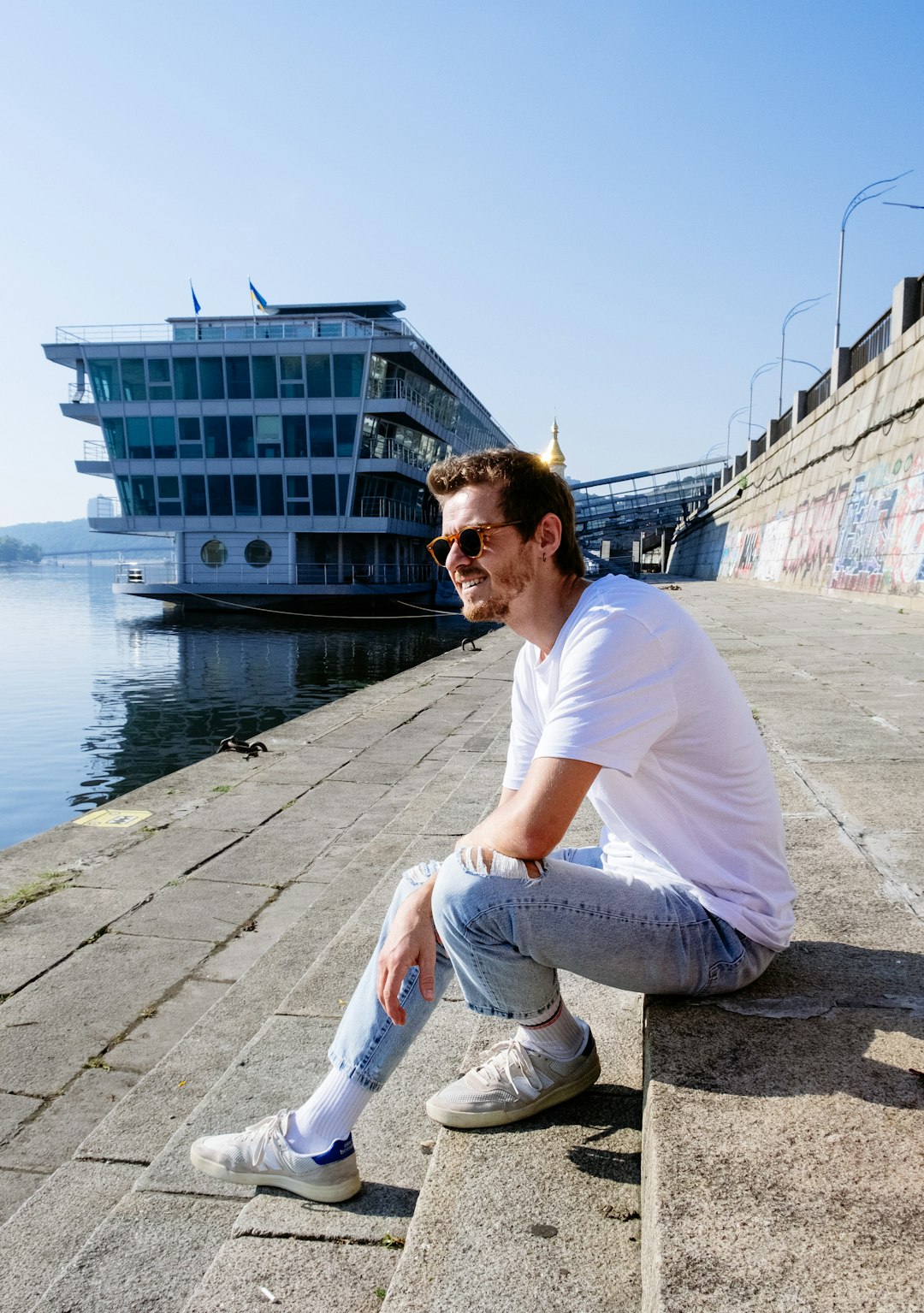 man in white t-shirt and gray denim jeans sitting on concrete dock during daytime
