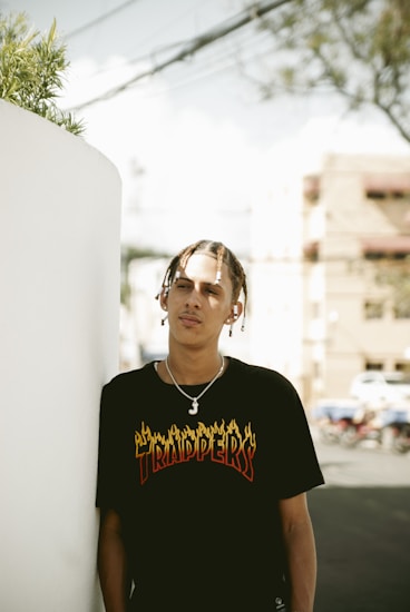 A person with braided hair stands outdoors, leaning against a white wall. They are wearing a black t-shirt with graphic text that reads 'TRAPPERS'. The background features blurred urban elements, such as buildings and a street.