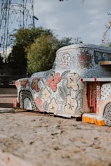 A mosaic art installation shaped like a vehicle is adorned with intricate tile patterns featuring flowers, skulls, and hearts. The vehicle is parked on a dirt surface with trees and a metal structure in the background. The mosaic art incorporates a variety of colors including red, black, and white tiles.