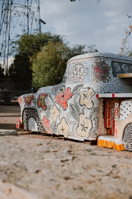 A mosaic art installation shaped like a vehicle is adorned with intricate tile patterns featuring flowers, skulls, and hearts. The vehicle is parked on a dirt surface with trees and a metal structure in the background. The mosaic art incorporates a variety of colors including red, black, and white tiles.