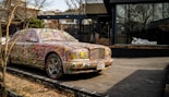 A car parked on the side of a road is covered in colorful, abstract paint splatters, making it a striking visual piece of art. Behind the car, there is a modern building featuring large glass windows. A white statue of a reclining figure is visible through the window, adding an additional element of interest.