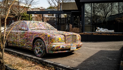 A car parked on the side of a road is covered in colorful, abstract paint splatters, making it a striking visual piece of art. Behind the car, there is a modern building featuring large glass windows. A white statue of a reclining figure is visible through the window, adding an additional element of interest.