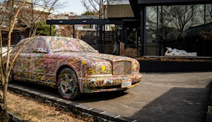 A car parked on the side of a road is covered in colorful, abstract paint splatters, making it a striking visual piece of art. Behind the car, there is a modern building featuring large glass windows. A white statue of a reclining figure is visible through the window, adding an additional element of interest.