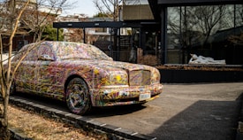 A car parked on the side of a road is covered in colorful, abstract paint splatters, making it a striking visual piece of art. Behind the car, there is a modern building featuring large glass windows. A white statue of a reclining figure is visible through the window, adding an additional element of interest.