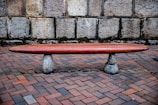 A minimalist wooden bench rests on two stone supports, situated on a brick-paved ground with a stone wall as the backdrop. The bench is elongated and has a sleek, polished finish, contrasting with the rough texture of the stones and brick.