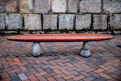 A minimalist wooden bench rests on two stone supports, situated on a brick-paved ground with a stone wall as the backdrop. The bench is elongated and has a sleek, polished finish, contrasting with the rough texture of the stones and brick.