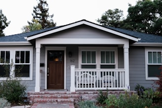 A small, single-story house with gray siding and a brick foundation features a wooden front door and a white porch railing. Two chairs are placed on the porch, and there are three windows with white trim. The yard is landscaped with shrubs and flowering plants, and tall trees are visible in the background.