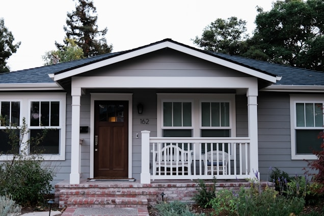 A small, single-story house with gray siding and a brick foundation features a wooden front door and a white porch railing. Two chairs are placed on the porch, and there are three windows with white trim. The yard is landscaped with shrubs and flowering plants, and tall trees are visible in the background.
