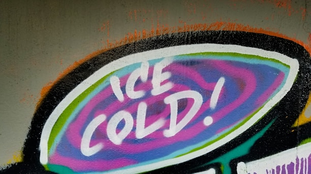Graffiti artwork featuring the words 'ICE COLD!' in bold, white letters. The text is set against a vibrant, swirling background of purple, blue, and pink hues encased in a black and white outline.