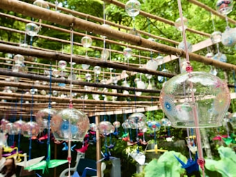 Numerous glass wind chimes are suspended from wooden beams in an outdoor setting. The chimes have intricate illustrated patterns and are surrounded by vibrant paper cranes. Bright green foliage is visible in the background, giving a natural and serene atmosphere.