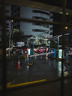 A nighttime view of a parking area with wet pavements reflecting street lights. Several cars, including a red one in focus, are waiting at a barrier gate. Dim lighting gives a subdued, urban atmosphere. The background features a large office building with illuminated signage.