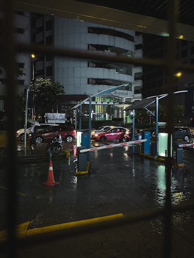 A nighttime view of a parking area with wet pavements reflecting street lights. Several cars, including a red one in focus, are waiting at a barrier gate. Dim lighting gives a subdued, urban atmosphere. The background features a large office building with illuminated signage.
