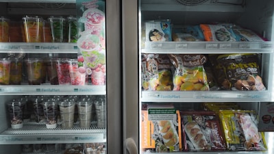 A refrigerated display case containing a variety of packaged goods. The left side features colorful beverage cups and a promotional sign for a watermelon lychee frappe. The right side has shelves with packaged snacks such as cheese and mochi items. Pricing labels are visible above each shelf.