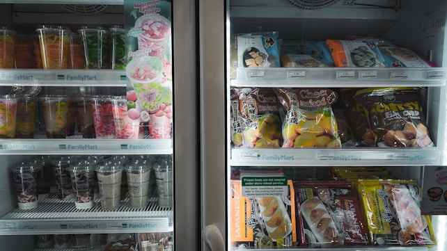 A refrigerated display case containing a variety of packaged goods. The left side features colorful beverage cups and a promotional sign for a watermelon lychee frappe. The right side has shelves with packaged snacks such as cheese and mochi items. Pricing labels are visible above each shelf.