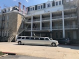 A long white limousine is parked in front of a large, historic-looking stone building with several stories. The building has white railings and multiple windows, and there is an external staircase on the side. The sky is clear with a few clouds.