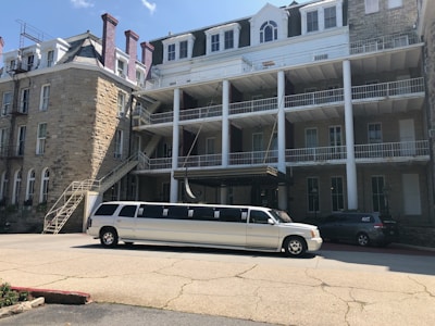 A long white limousine is parked in front of a large, historic-looking stone building with several stories. The building has white railings and multiple windows, and there is an external staircase on the side. The sky is clear with a few clouds.