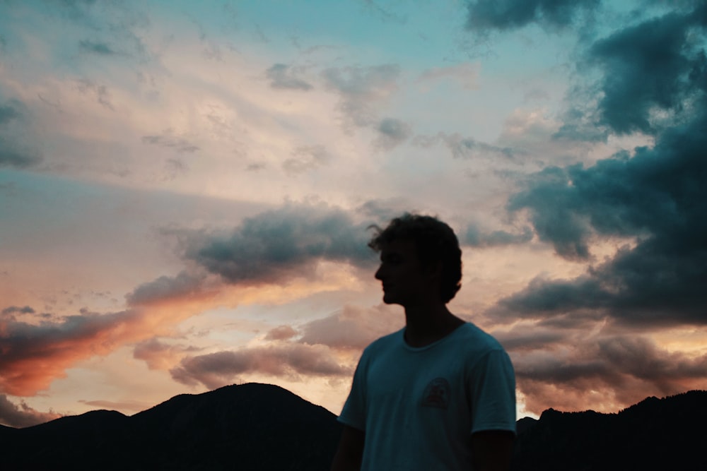 man in white crew neck t-shirt standing on mountain under white clouds during daytime