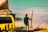 A person stands near the edge of a cliff holding a surfboard, overlooking a vast expanse of clouds. Next to the person is a vintage van with a rooftop tent, featuring colorful artwork and text. A small folding table with various items is set up beside the van.