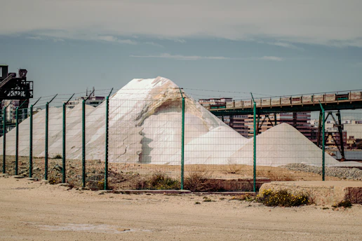 A large white pile of salt or similar material is situated behind a green wire fence. An industrial structure with metal beams and a conveyor belt runs above the mound. In the background, there are buildings and a clear blue sky.