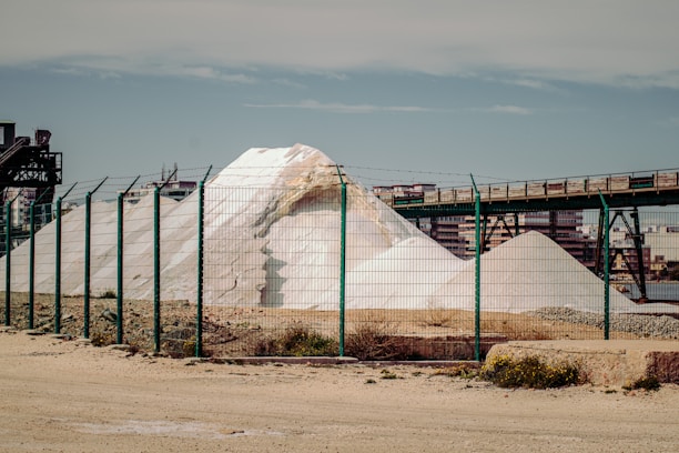 A large white pile of salt or similar material is situated behind a green wire fence. An industrial structure with metal beams and a conveyor belt runs above the mound. In the background, there are buildings and a clear blue sky.