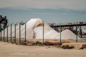 A large white pile of salt or similar material is situated behind a green wire fence. An industrial structure with metal beams and a conveyor belt runs above the mound. In the background, there are buildings and a clear blue sky.