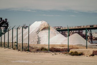 A large white pile of salt or similar material is situated behind a green wire fence. An industrial structure with metal beams and a conveyor belt runs above the mound. In the background, there are buildings and a clear blue sky.