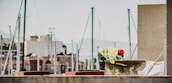 A construction worker wearing a fluorescent safety jacket and red helmet is working near a concrete structure. In the background, there are several unfocused yacht masts and indistinct buildings, suggesting a marine or port environment.
