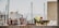 A construction worker wearing a fluorescent safety jacket and red helmet is working near a concrete structure. In the background, there are several unfocused yacht masts and indistinct buildings, suggesting a marine or port environment.