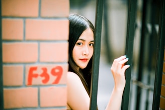 A young woman with long dark hair is looking through a black iron fence. Her hand is gently holding one of the bars. The background features a brick wall with 'F39' painted in red.