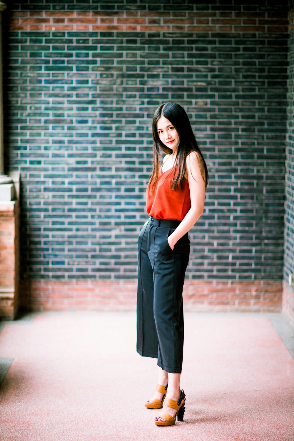 woman in red tank top and black pants standing beside brick wall