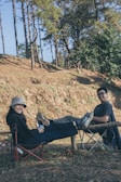 Two people are sitting outdoors in foldable camping chairs, smiling and relaxing with their legs propped up on a wooden log. They are surrounded by trees and grass, suggesting a serene, natural setting. The person on the left is wearing a bucket hat and holding a mug, while the person on the right is casually dressed in a t-shirt.