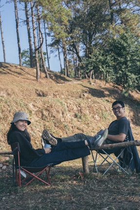 Two people are sitting outdoors in foldable camping chairs, smiling and relaxing with their legs propped up on a wooden log. They are surrounded by trees and grass, suggesting a serene, natural setting. The person on the left is wearing a bucket hat and holding a mug, while the person on the right is casually dressed in a t-shirt.