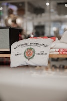 A folded t-shirt with a printed design featuring the text 'Rosemont Barbershop & Grooming Supply' alongside a floral emblem. The establishment details 'Est. Denver 2019 Colorado' are also visible on the shirt. It is displayed on a shelf in a retail environment with a blurred background.