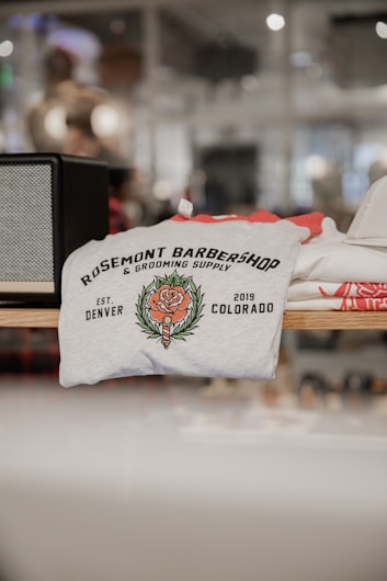 A folded t-shirt with a printed design featuring the text 'Rosemont Barbershop & Grooming Supply' alongside a floral emblem. The establishment details 'Est. Denver 2019 Colorado' are also visible on the shirt. It is displayed on a shelf in a retail environment with a blurred background.
