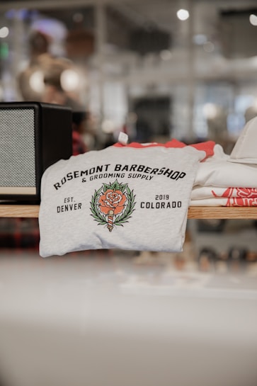 A folded t-shirt with a printed design featuring the text 'Rosemont Barbershop & Grooming Supply' alongside a floral emblem. The establishment details 'Est. Denver 2019 Colorado' are also visible on the shirt. It is displayed on a shelf in a retail environment with a blurred background.