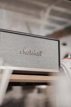 A vintage-style Marshall speaker with a textured front grille and the logo prominently displayed in gold lettering. The speaker is placed on a wooden surface in a minimalist setting.