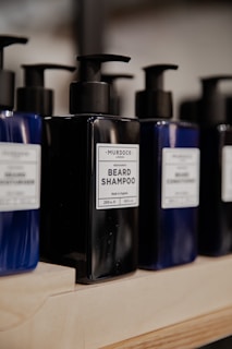 Several bottles of beard shampoo and conditioner with black pump dispensers are displayed on a wooden shelf. The central bottle is labeled 'Murdock London Beard Shampoo.' The bottles are dark blue and black, creating a sleek and modern look.