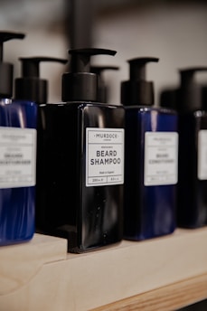 Several bottles of beard shampoo and conditioner with black pump dispensers are displayed on a wooden shelf. The central bottle is labeled 'Murdock London Beard Shampoo.' The bottles are dark blue and black, creating a sleek and modern look.
