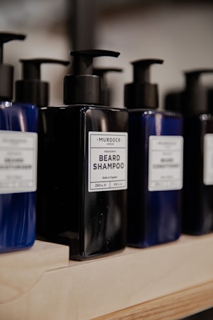 Several bottles of beard shampoo and conditioner with black pump dispensers are displayed on a wooden shelf. The central bottle is labeled 'Murdock London Beard Shampoo.' The bottles are dark blue and black, creating a sleek and modern look.