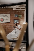A barber wearing a mask is cutting a person's hair inside a barber shop. The interior features exposed brick walls with framed signs and a plant hanging on the wall. The person getting a haircut is seated in a barber chair and draped with a striped cover. The atmosphere is cozy and warm, highlighted by soft lighting.