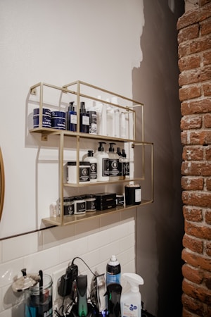 A wall-mounted metal shelf holds an assortment of grooming and hair care products, including bottles and jars with labels. Below the shelf, various electronic grooming tools and bottles, such as spray bottles, are placed on a counter. The background features a mix of white subway tiles and a partially painted wall, alongside a brick wall.