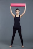 A person stands with legs apart and raises a rolled yoga mat overhead. They wear a black tank top and black leggings, appearing to be prepared for a fitness activity. The background is a simple, solid gray color.