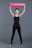 A person stands with legs apart and raises a rolled yoga mat overhead. They wear a black tank top and black leggings, appearing to be prepared for a fitness activity. The background is a simple, solid gray color.