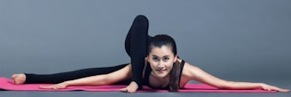 A woman is performing a flexible yoga pose on a pink yoga mat, extending one leg back and the other forward, with her arms stretched out in front. She is wearing a black outfit against a plain gray background.
