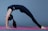 A person performing a yoga backbend pose on a pink yoga mat, wearing a black outfit against a gray background.