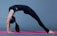 A person performing a yoga backbend pose on a pink yoga mat, wearing a black outfit against a gray background.