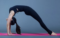 A person performing a yoga backbend pose on a pink yoga mat, wearing a black outfit against a gray background.