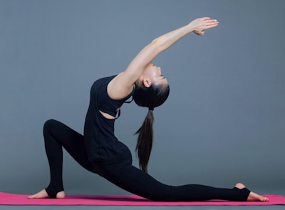 A person is performing a deep lunge yoga pose on a pink mat, with arms extended overhead. The background is plain and gray, while the athlete wears a fitted black outfit.