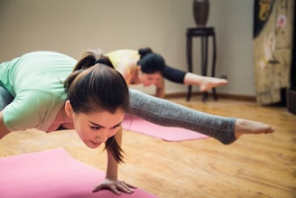 Two people are practicing yoga indoors, performing a challenging balancing pose on yoga mats. They are positioned on a wooden floor in a well-lit room, wearing athletic clothing. The focus is on the person in front who has their hair tied back and is balancing skillfully.